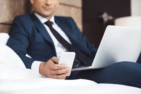 Zakenman op bed met telefoon en laptop — Stockfoto