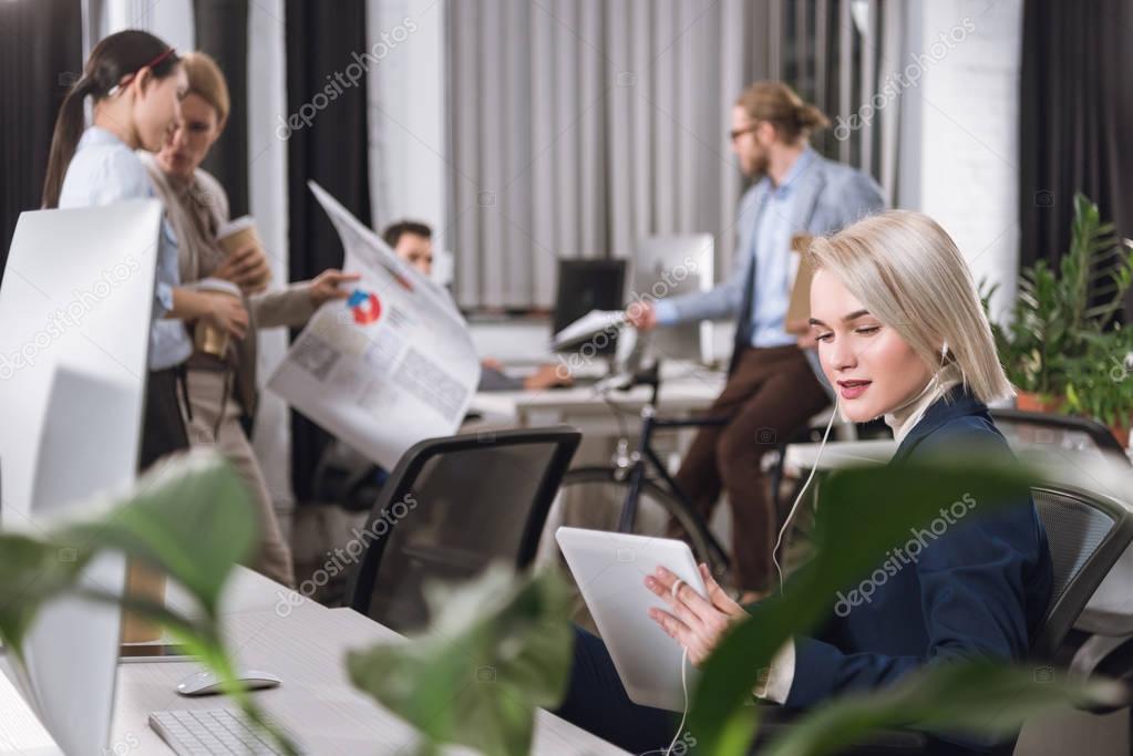 Selective focus of businesswoman in earphones using tablet at workplace in office