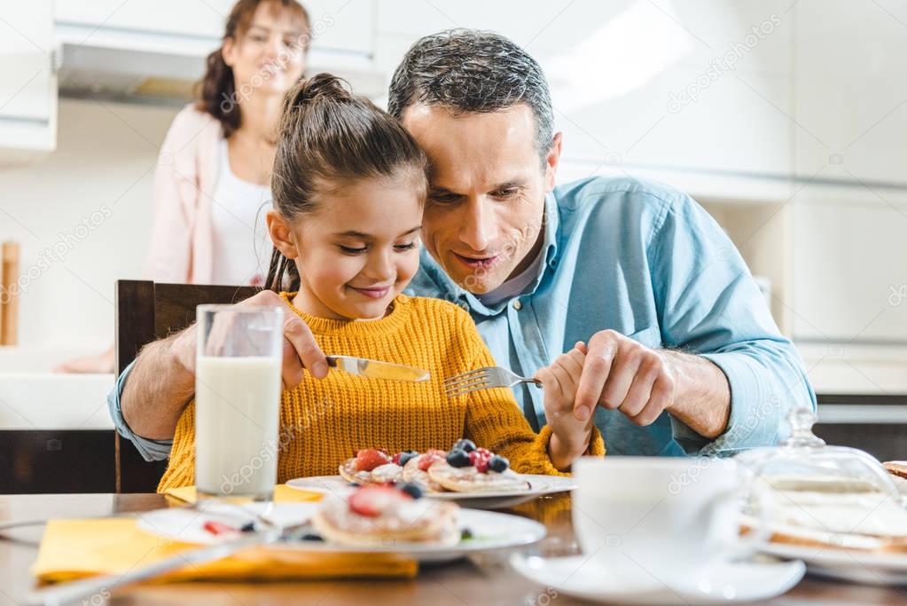 cheerful family together eating pancakes with berries at table on kitchen