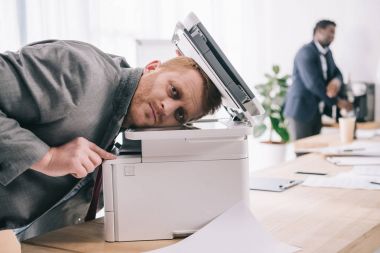 overworked young businessman leaning on copier at office clipart