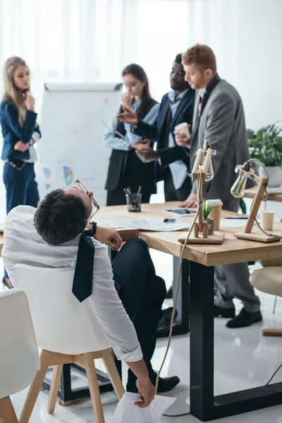 年轻的工作过度的生意人睡在会议室 而同事交谈 — 图库照片