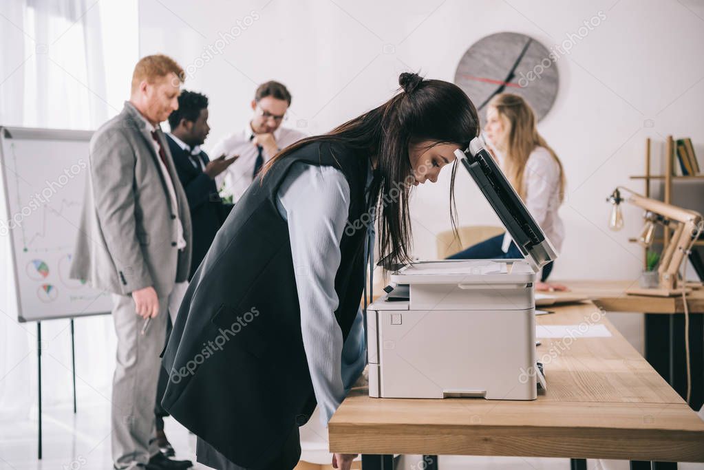 exhausted sleepy businesswoman leaning on copier at office while colleagues having conversation