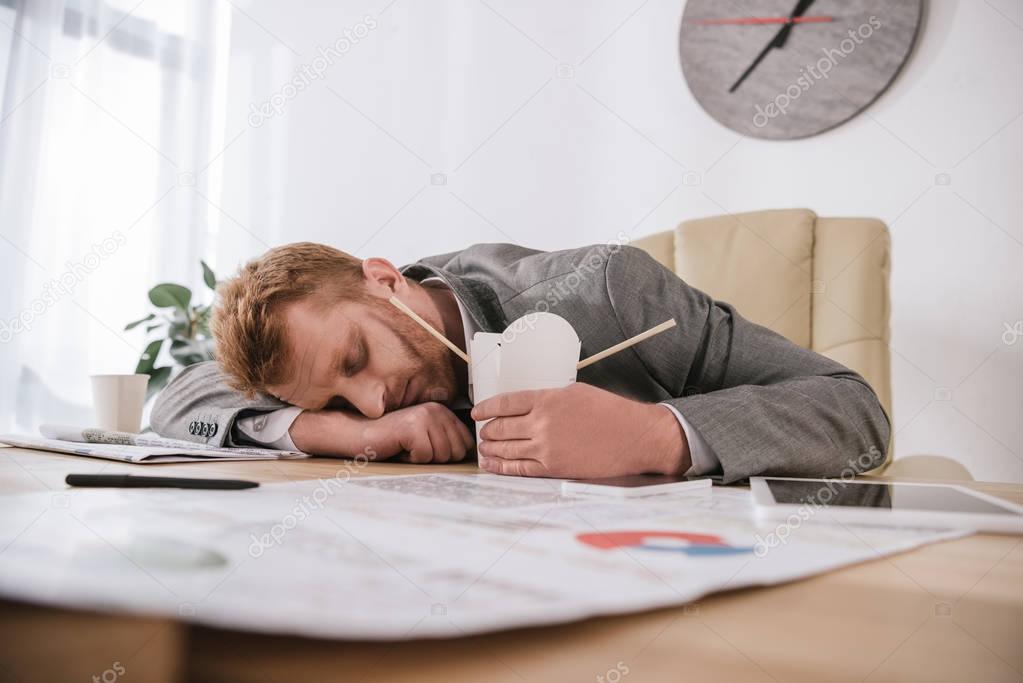 exhausted young businessman sleeping with box of take away noodles at workplace