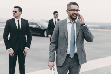 smiling businessman walking with bodyguards and talking by smartphone  clipart