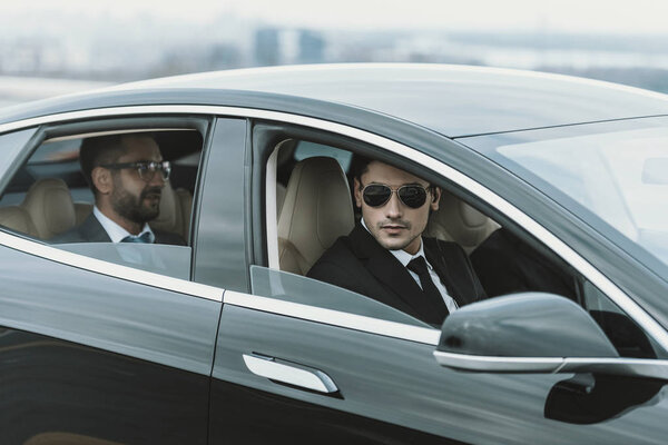 два телохранителя и бизнесмен в очках, сидящие в черной машине
 