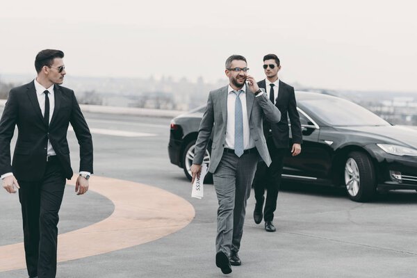 businessman walking with bodyguards on helipad and talking by smartphone 