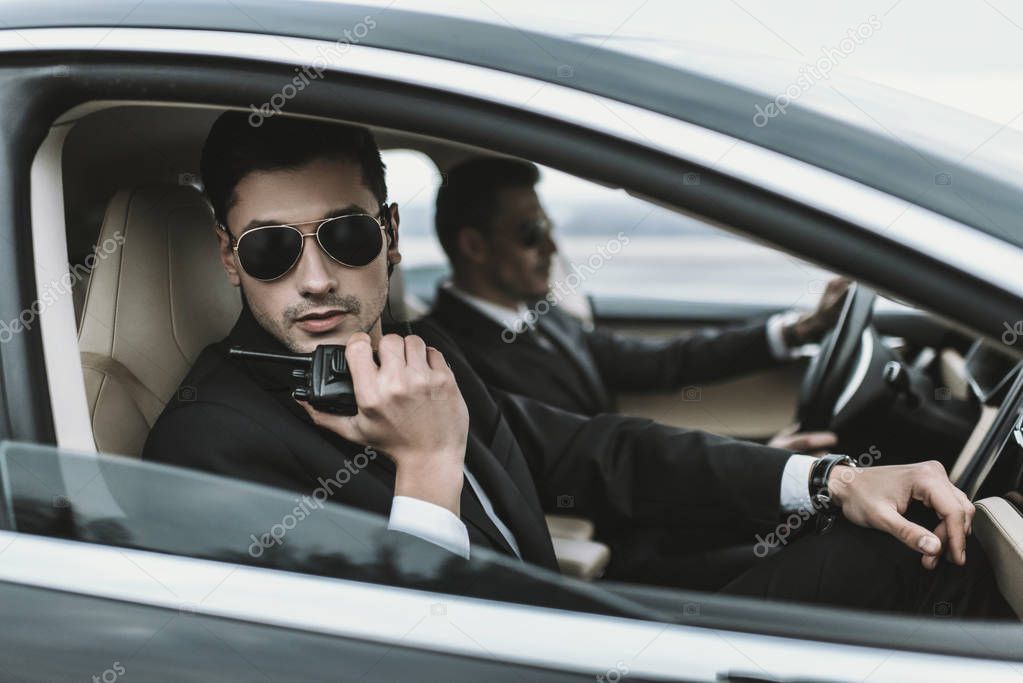 bodyguard in sunglasses talking by portable radio while sitting in car