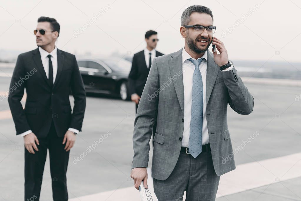 smiling businessman walking with bodyguards and talking by smartphone 