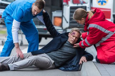 young male and female paramedics helping injured man on the street clipart