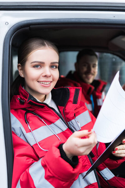 smiling young female paramedic holding clipboard and looking at camera