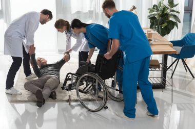doctors helping fell mature man to sit on wheelchair clipart