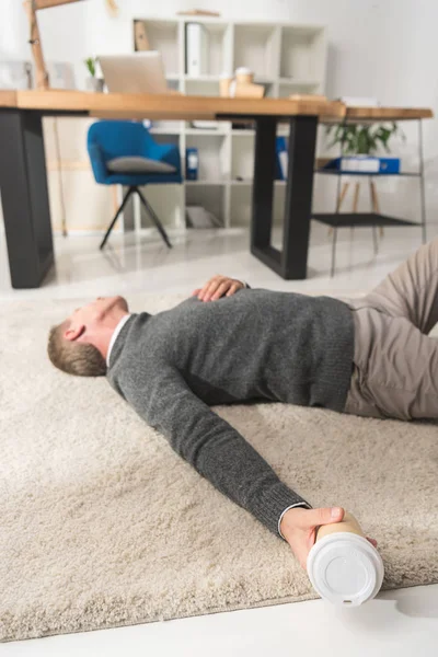 オフィスの床の上に横たわると 使い捨てのコーヒー カップを手で保持している無意識の男 — ストック写真