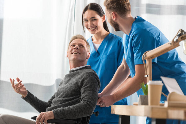 улыбающиеся врачи и пациент на инвалидной коляске разговаривают в больнице
