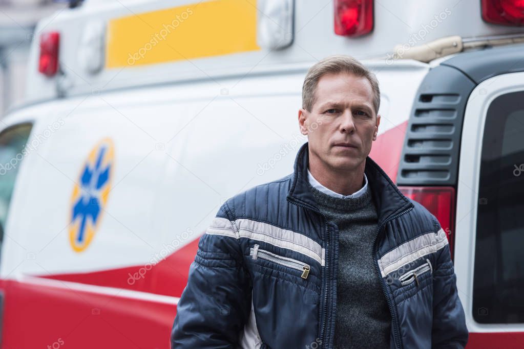 paramedic in casual clothes standing in front of ambulance and looking at camera