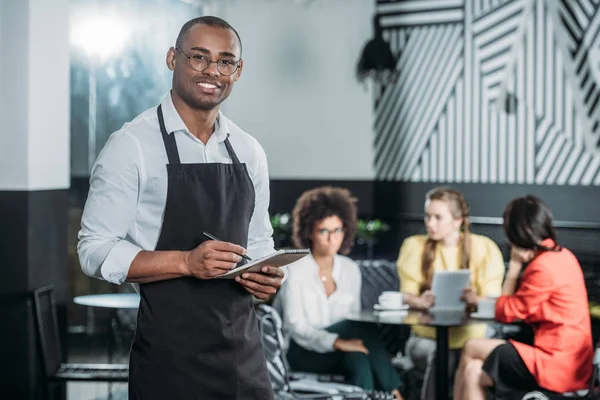 happy young waiter writing in notepad with multiethnic group of women sitting blurred on background