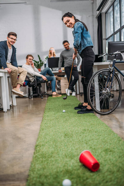 счастливая женщина играет в мини-гольф с коллегами в современном офисе
