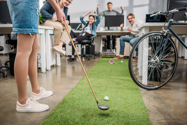 обрезанный образ девушки, играющей в мини-гольф с коллегами в современном офисе
 