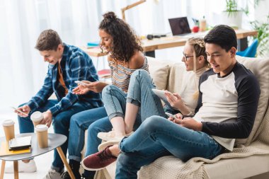 happy multicultural teens sitting on sofa with digital devices  clipart