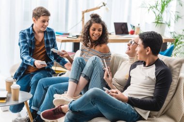 smiling multiethnic teenagers sitting on sofa with gadgets 