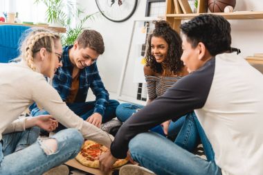 multiethnic teen friends sitting on floor and eating pizza clipart