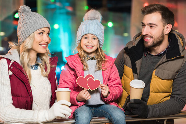 happy parents with coffee to go looking at cute little daughter holding hearts symbol on rink