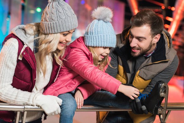 smiling young parents looking at adorable little daughter wearing skates on rink 