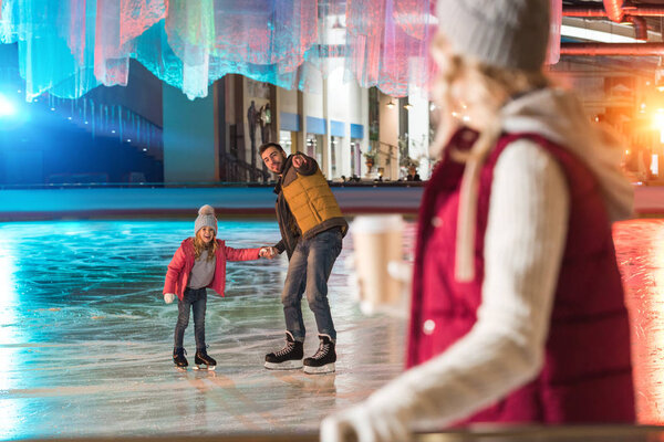 счастливые отец и дочь держась за руки и указывая пальцем на молодую женщину, пьющую кофе на переднем плане
