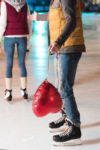 частичный вид молодого человека, держащего воздушный шар в форме сердца, в то время как девушка уходит на каток
