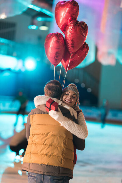 счастливая молодая пара с подарочной коробкой и шарами в форме сердца, обнимающие на катке
 