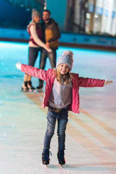 прелестная маленькая девочка, стоящая с распростертыми объятиями и улыбающаяся в камеру, пока родители стоят позади на катке
