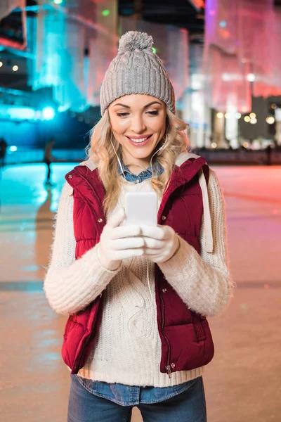 Ελκυστική Χαμογελώντας Νεαρή Γυναίκα Στα Ακουστικά Χρησιμοποιώντας Smartphone Στο Παγοδρόμιο — Φωτογραφία Αρχείου