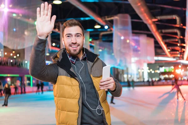 イヤホン スマート フォンを押し スケート場の中に立って手を振って笑顔の若い男 — ストック写真