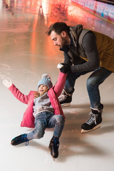 Отец держит за руку милую маленькую дочь, падающую на каток
