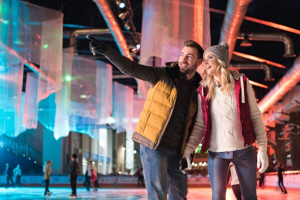 счастливая молодая пара в теплой одежде, держась за руки и глядя на каток
