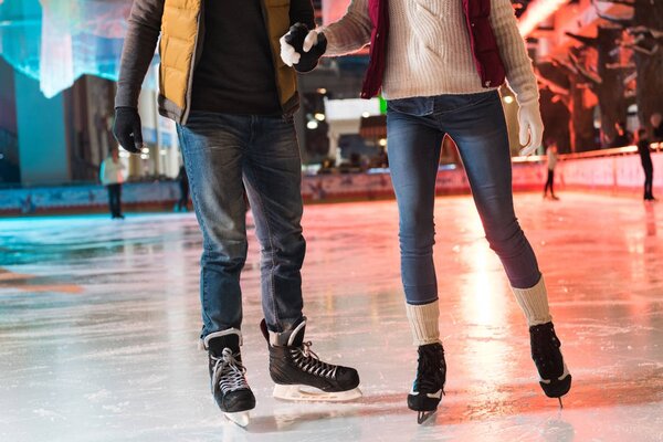 cropped shot of young couple in skates holding hands and ice skating on rink