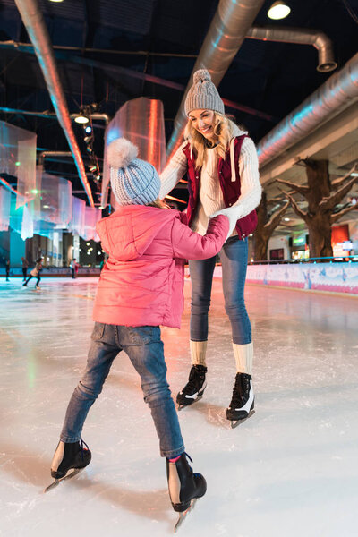 happy mother and daughter holding hands and looking at each other on skating rink 