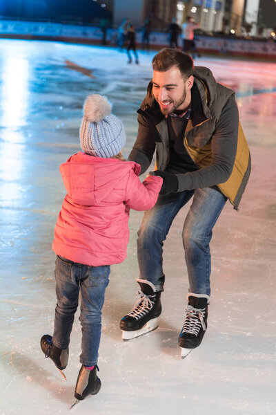 счастливые отец и дочь держатся за руки и смотрят друг на друга на катке
 