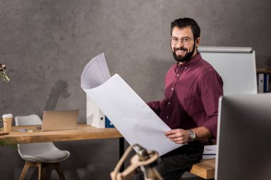 Blueprint tutarak ve kameraya bakarak gülümseyen mimar