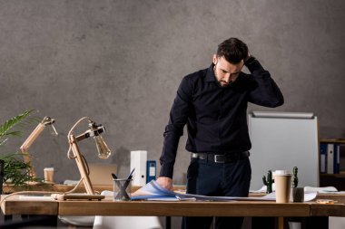 thoughtful architect standing near working table and looking at blueprints clipart