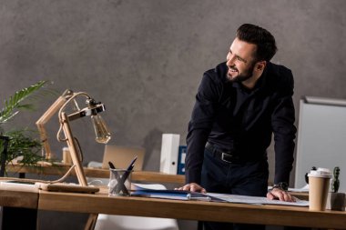 smiling architect leaning on working table and looking away clipart