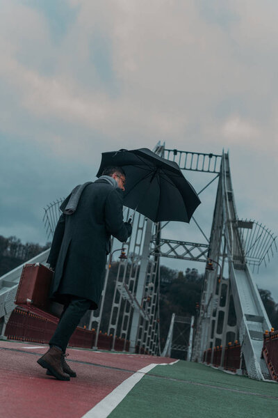 вид снизу на красивого мужчину средних лет с зонтиком и багажом, идущего по пешеходному мосту
