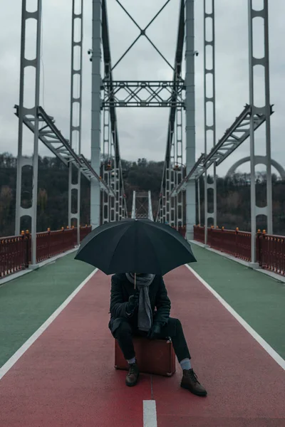 歩道橋でスーツケースの上に座って傘を持つスタイリッシュな男  — 無料ストックフォト