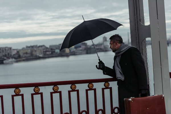 adult man with umbrella and luggage walking by bridge