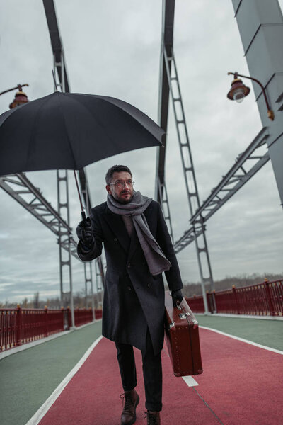 стильный взрослый мужчина с зонтиком и багажом ходьба по пешеходному мосту
