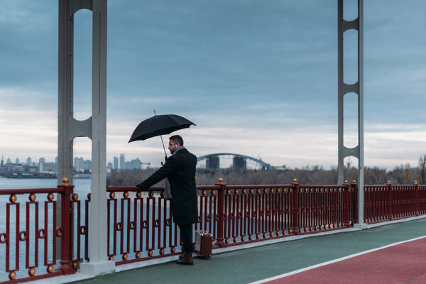 стильный одинокий мужчина с зонтиком и чемоданом, стоящий на мосту
