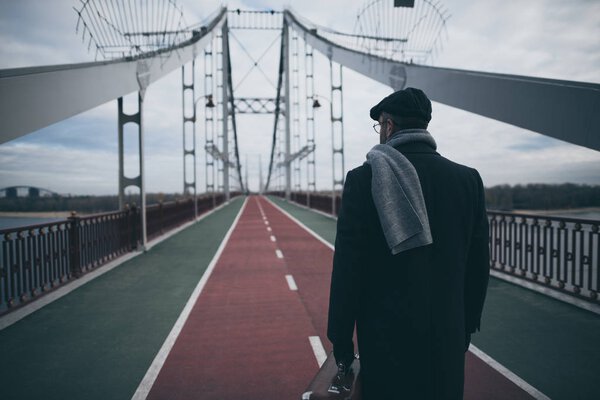 stylish man in coat and scarf with suitcase standing on pedestrian bridge