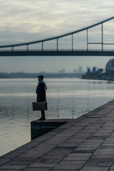 handsome adult man in coat with suitcase standing on river shore