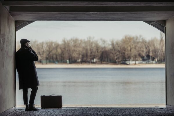 handsome adult man with suitcase smoking in tunnel in front of river shore