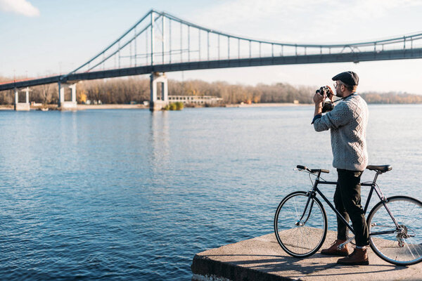 handsome man with vintage film camera and bike standing on river shore