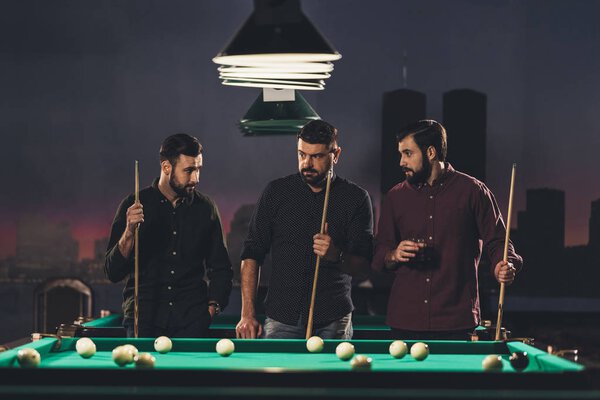 company of successful handsome men standing beside pool table with drink at bar